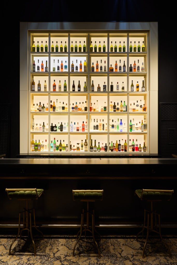 Backlit wall of liquor bottles arranged in a grid behind a bar, with chrome framing and bar stools in front.