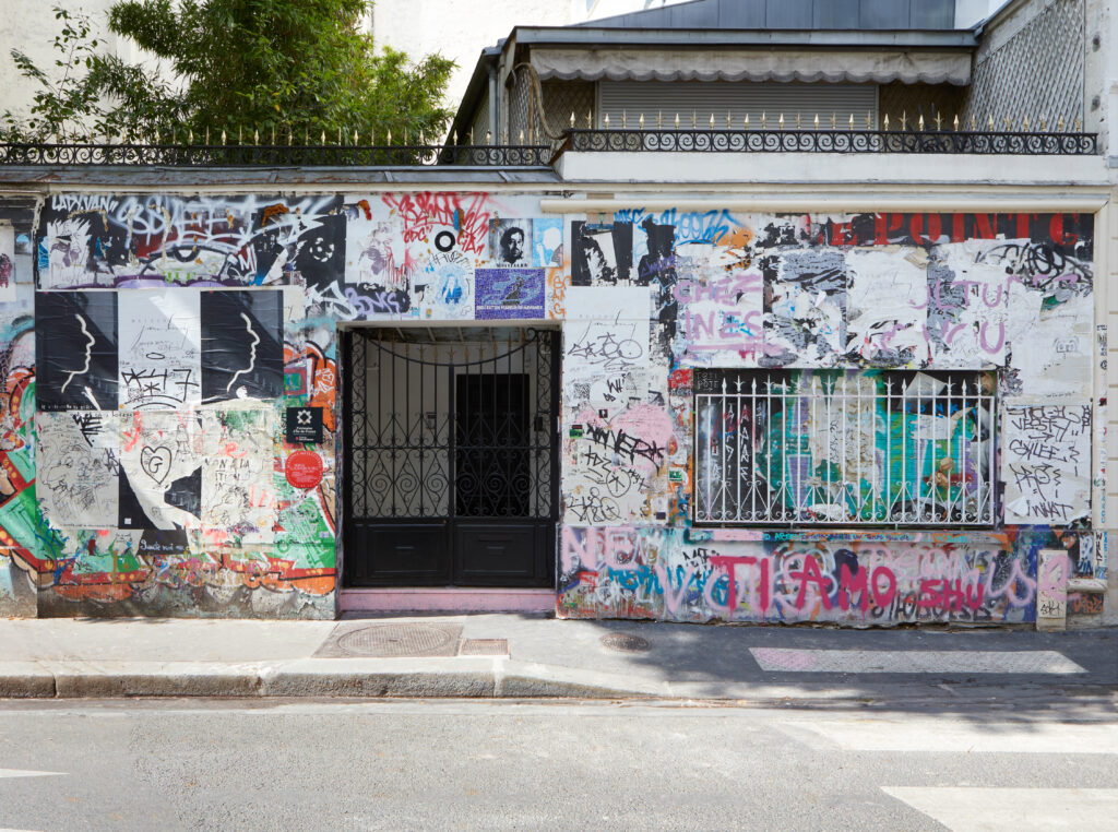 Wall and storefront covered in colorful graffiti with a black arched gate and a balcony above it on an urban street.