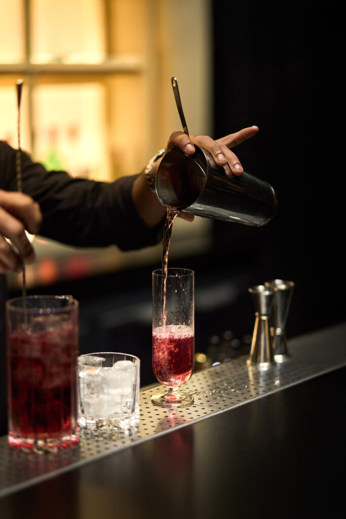 Bartender pouring a pink-red cocktail from a shaker into a tall glass at a bar.
