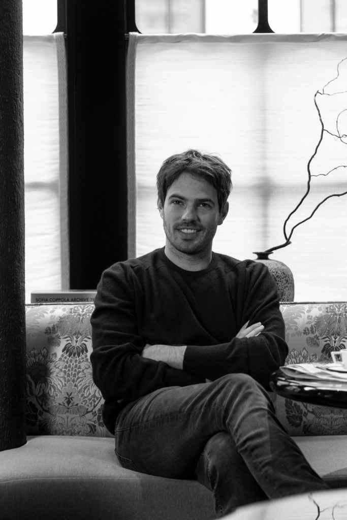 Man with short hair and light stubble sits on a patterned sofa with arms crossed, smiling slightly at the camera in a modern indoor setting.