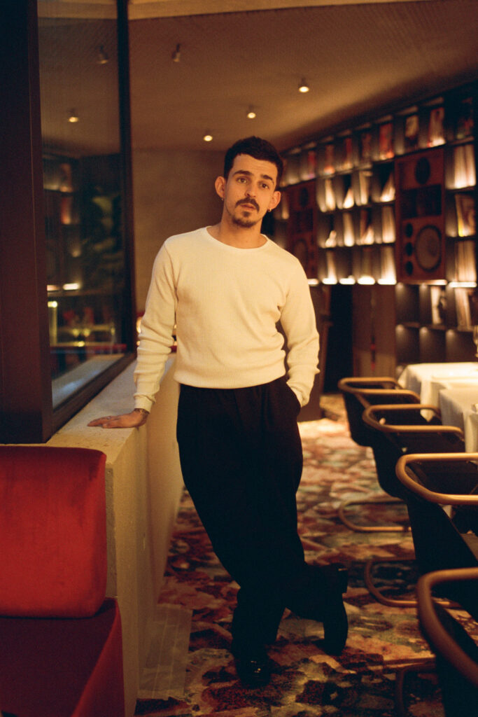 Man in a white sweater and black pants leans against a low wall in a warmly lit dining room.