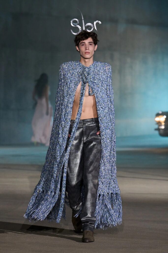 Runway model wearing a long blue patterned coat open at the front, showing metallic plaid pants, walking on a dimly lit stage.