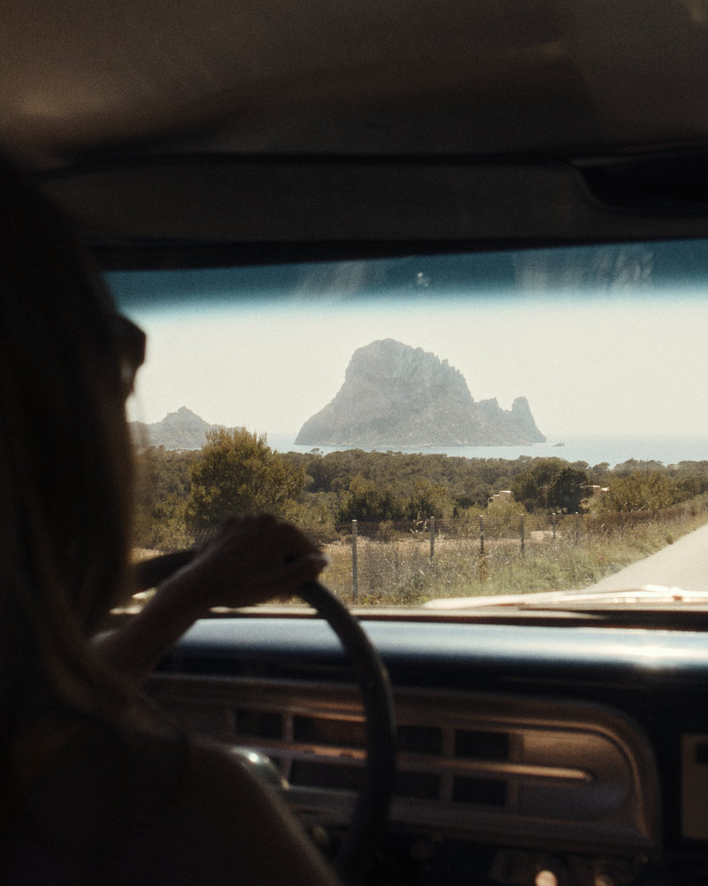 Driver's silhouette grips the steering wheel as a coastal landscape with a rocky island seen through the windshield.