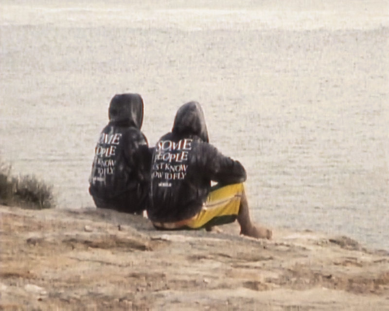 Two people wearing hoodies sit on a rocky shoreline, facing a calm body of water from behind.