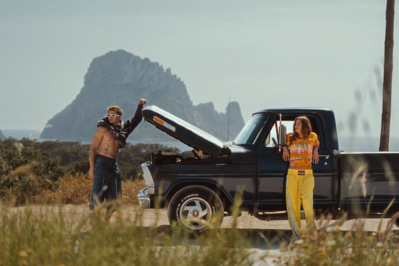 Shirtless man and a woman in yellow pants by a blue pickup with its hood raised, coastal rocks in the background.