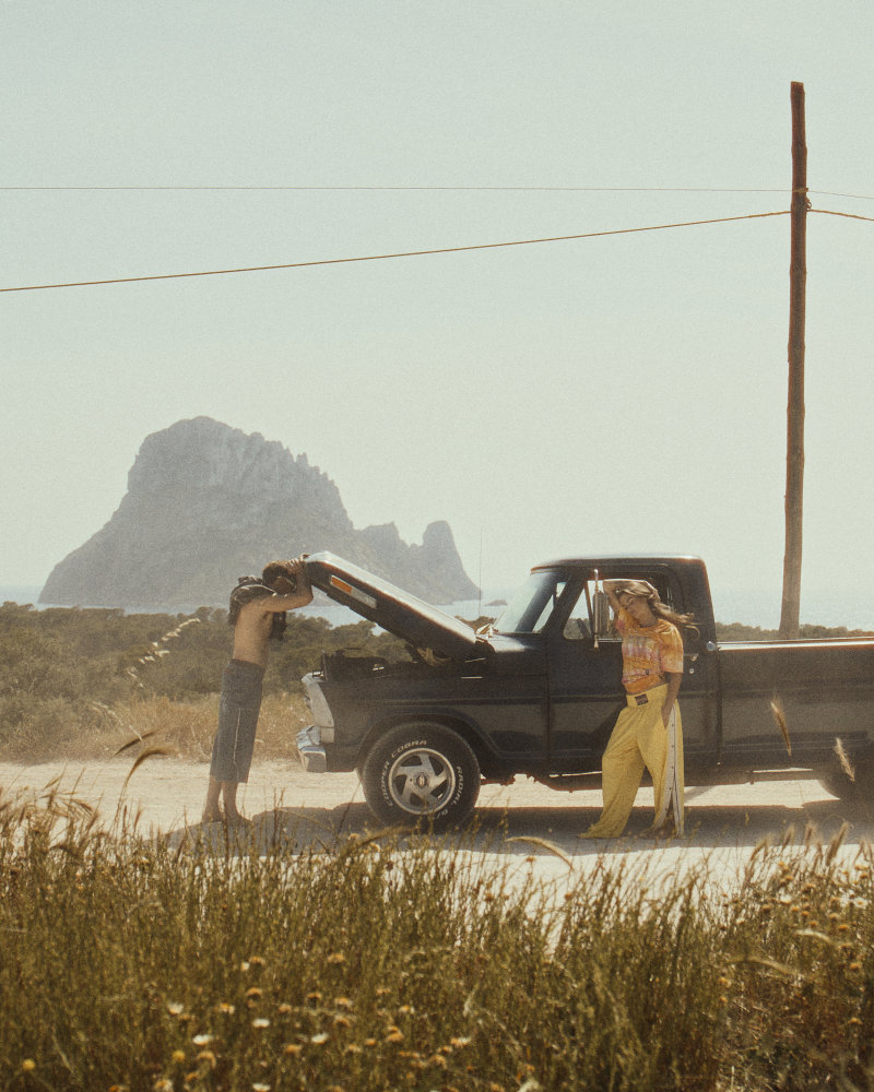 Two people by a black pickup on a dusty coastal road; one leans into the bed while the other stands nearby with a rocky backdrop in the distance.