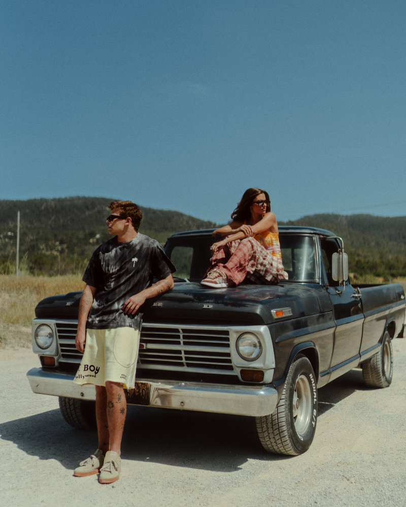 Two young adults sit on and lean against a vintage black pickup truck on a sunny rural road, with hills in the background.