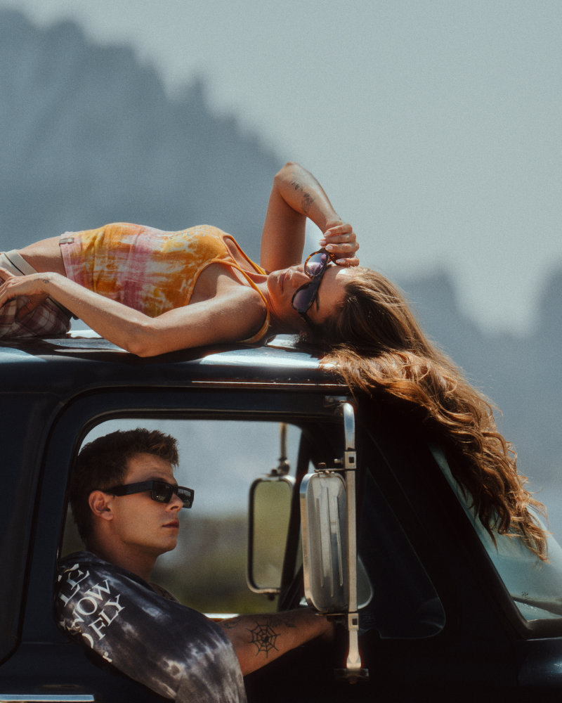Woman in an orange tank top lies on the roof of a black pickup, sunglasses on, while a man drives beside with sunglasses on.