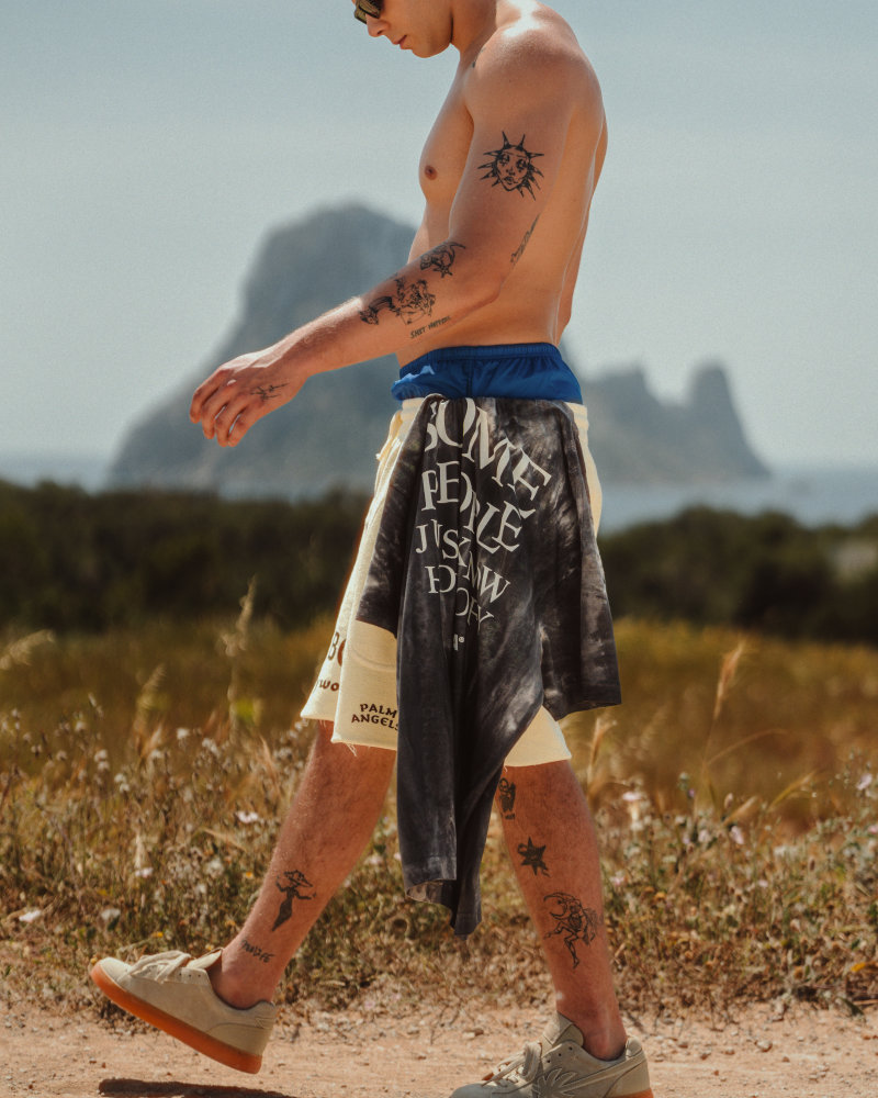 Young man shirtless in a sunny outdoor setting with tattoos on arms and legs, walking in shorts and sneakers, a towel draped around his waist; beach and rocky cliffs in the background.