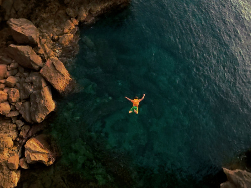 Aerial view of a person in an orange shirt jumping into turquoise water from a rocky cliff.