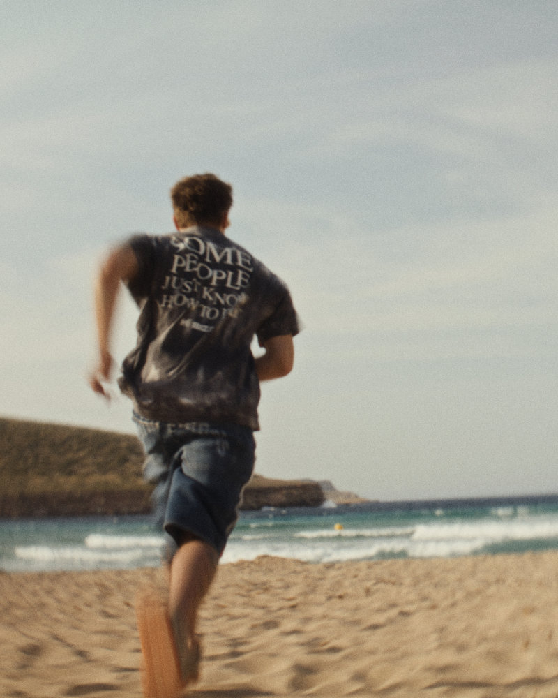 Person running on a sandy beach toward the ocean, wearing dark graphic T-shirt and shorts with waves in the background