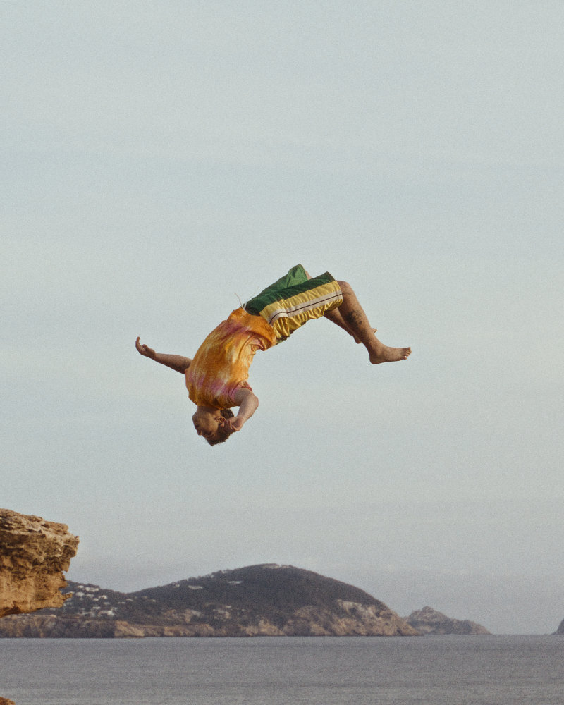 Person mid-air performing a backflip off a rocky cliff into the sea, wearing a tie-dye shirt and green-yellow shorts.