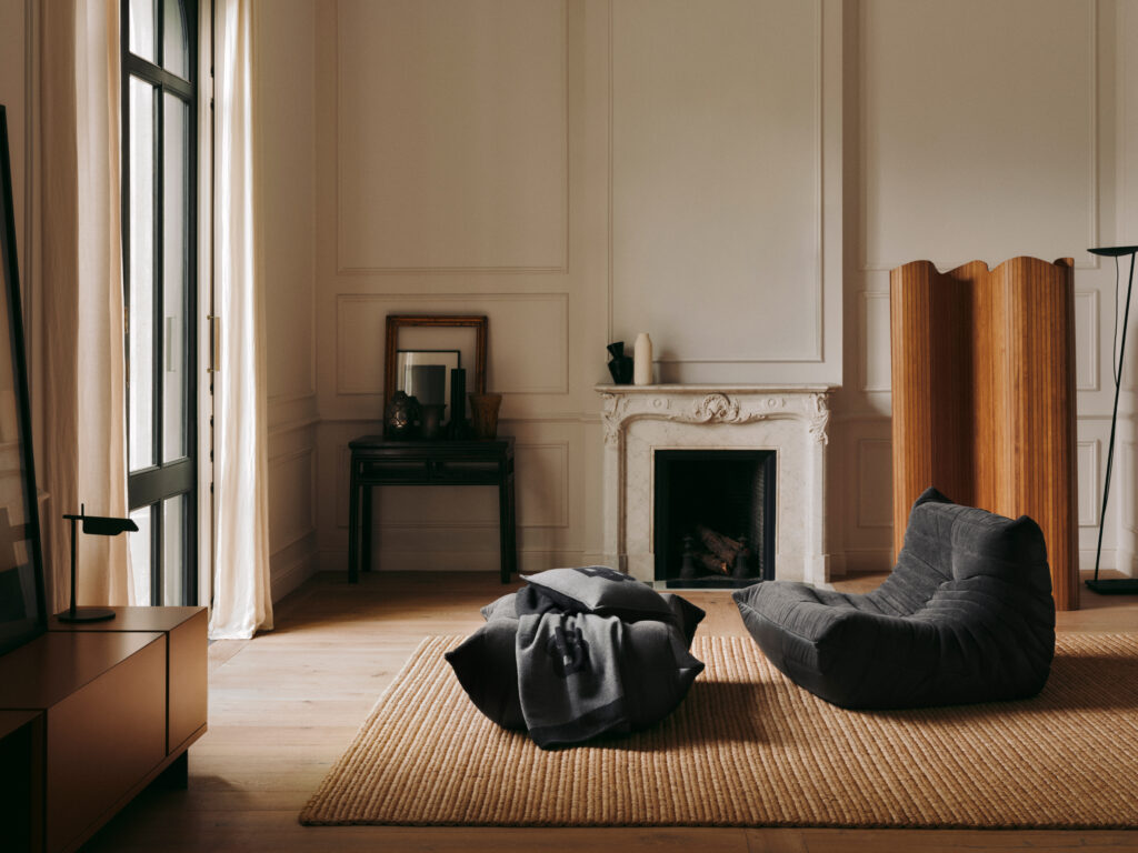 Cozy living room with beige paneled walls, a marble fireplace, and a wooden room divider bathed in natural light from tall windows.