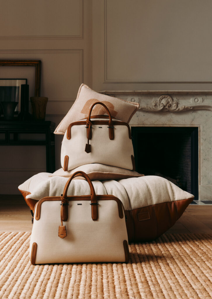 Beige suitcases with brown leather trims stacked on a cushion and rug in a stylish living room beside a fireplace mantel and pillow behind them.