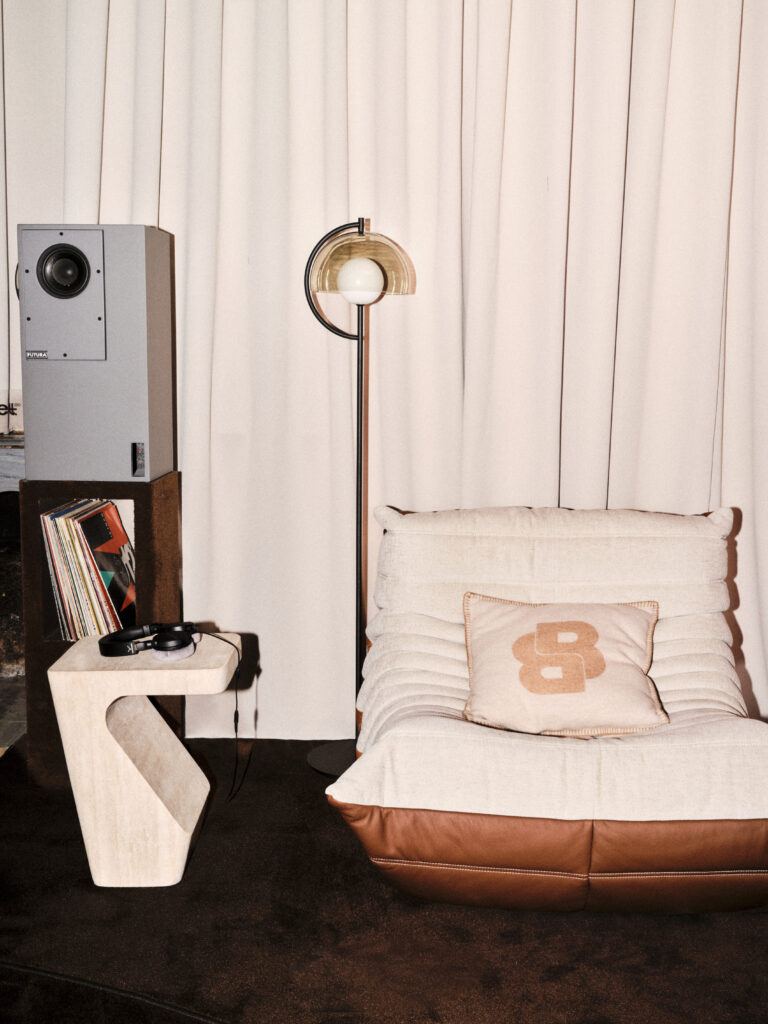 Mid-century living room corner with a tall gray speaker, a stylish arc-floor lamp, a beige cushioned chair, and a small rack of vinyl records with headphones on a white side table