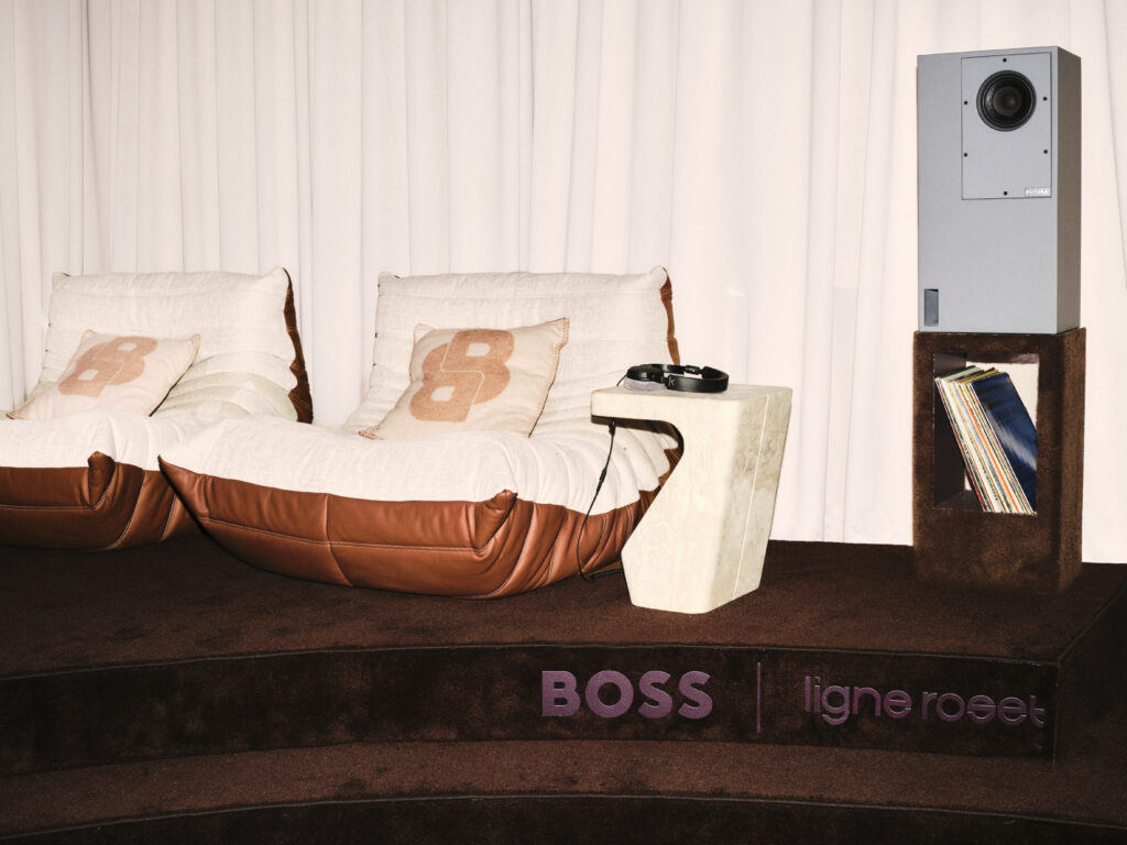 Cream sofa with beige cushions in a home showroom, a round side table with headphones, and a tall gray speaker on the right beside a bookshelf.