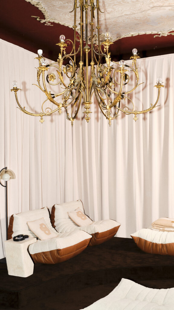 Gold ornate chandelier hanging from a ceiling above a dark brown sofa with cream and brown cushions, in a room with pale pink curtains.