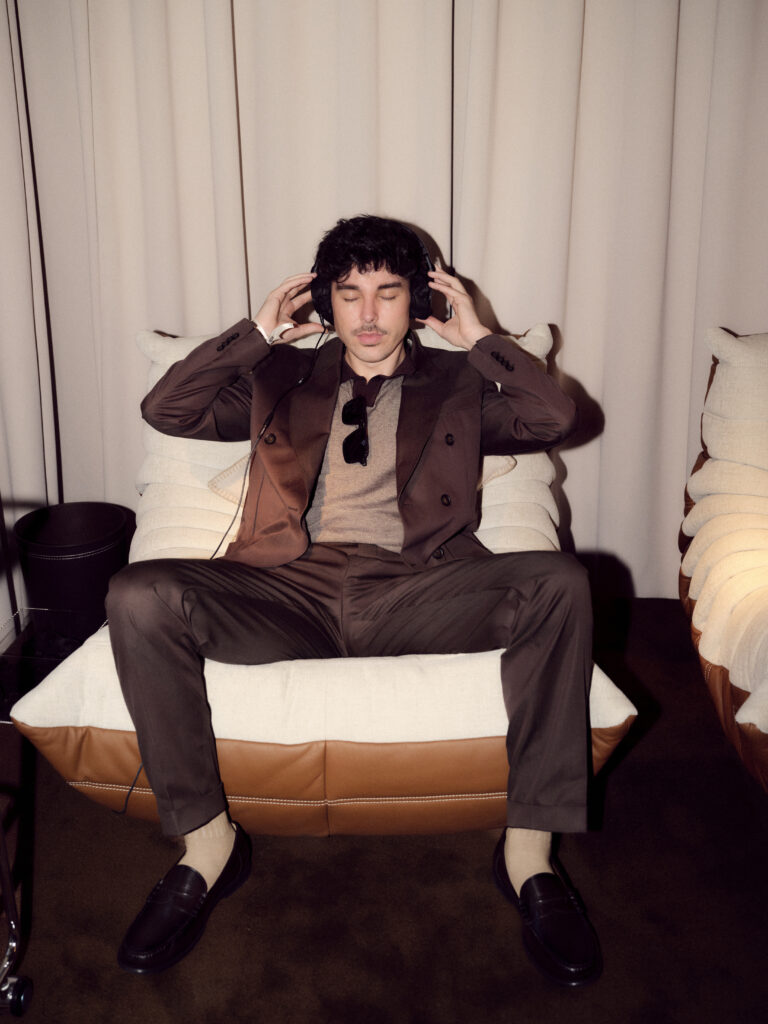 Man in a brown suit sits on a cream sofa with eyes closed, wearing large over-ear headphones—listening to music.