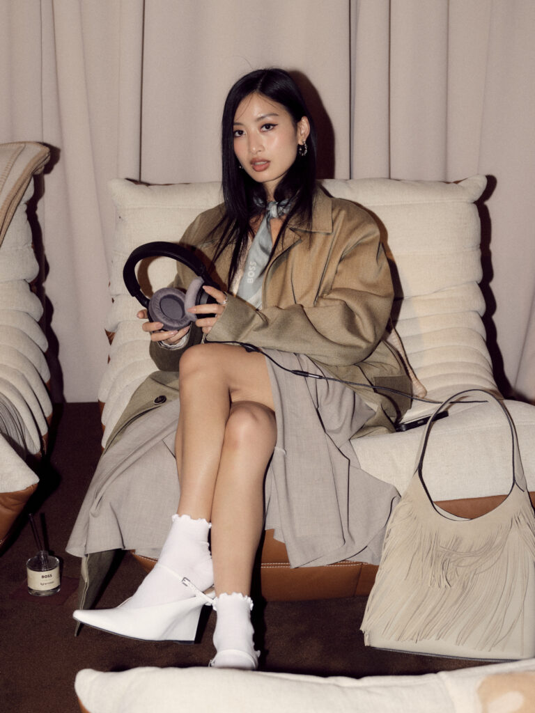 Young woman with straight black hair wearing a beige trench coat and light blue neck scarf sits on a cream sofa, holding gray over-ear headphones in front of her.