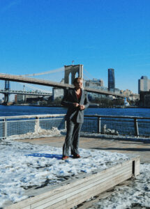 Jake Jensen in a suit standing on a snow-covered boardwalk with the Brooklyn Bridge and Manhattan skyline in the background.