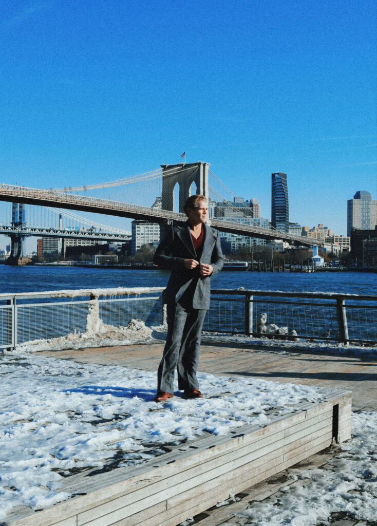 Jake Jensen in a suit standing on a snow-covered boardwalk with the Brooklyn Bridge and Manhattan skyline in the background.