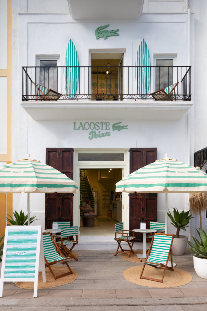 White Lacoste Ibiza storefront with an open door, balcony above, teal striped surfboards, and outdoor seating with green-and-white striped umbrellas.