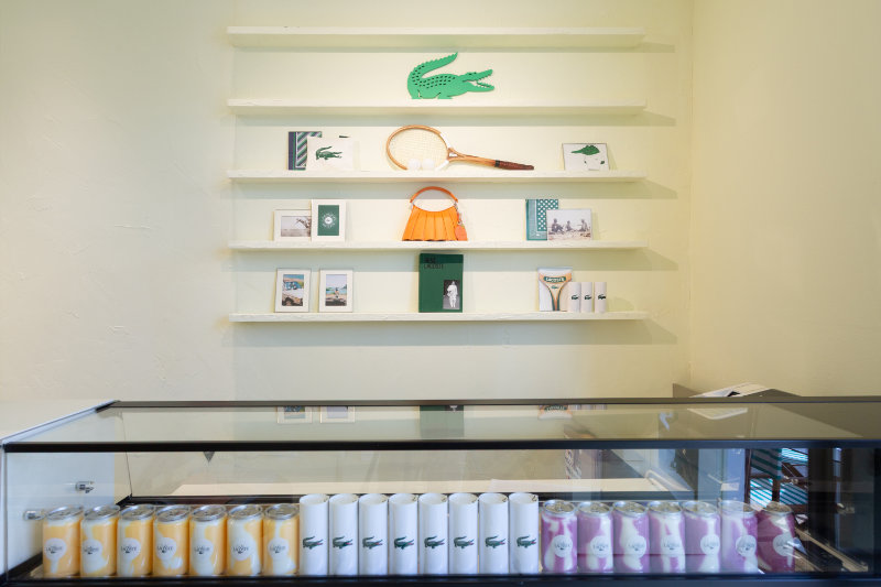 Boutique display on a pale wall with white shelves, a green crocodile logo, tennis racket, orange handbag, and small books; glass refrigerated counter below with pastel cans.