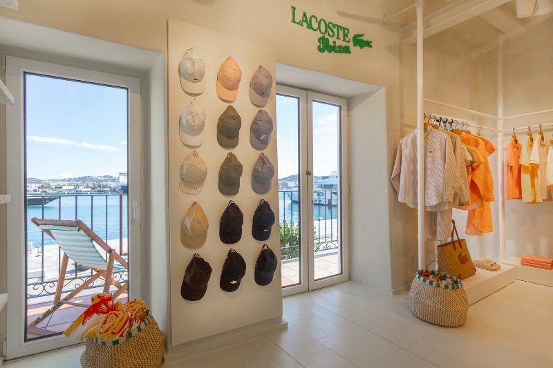 Bright shop interior with a wall of baseball caps beside glass doors and a Lacoste sign overhead.