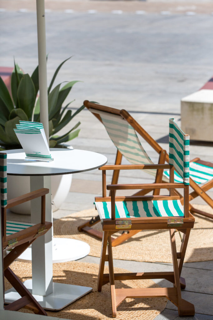 Outdoor patio scene with green-and-white striped deck chairs around a white round table, under a sunlit umbrella pole.