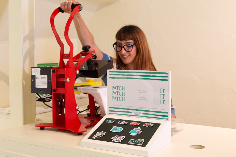 Woman with glasses operating a red heat-press/button press at a workstation, with sticker sheets and a patch display nearby