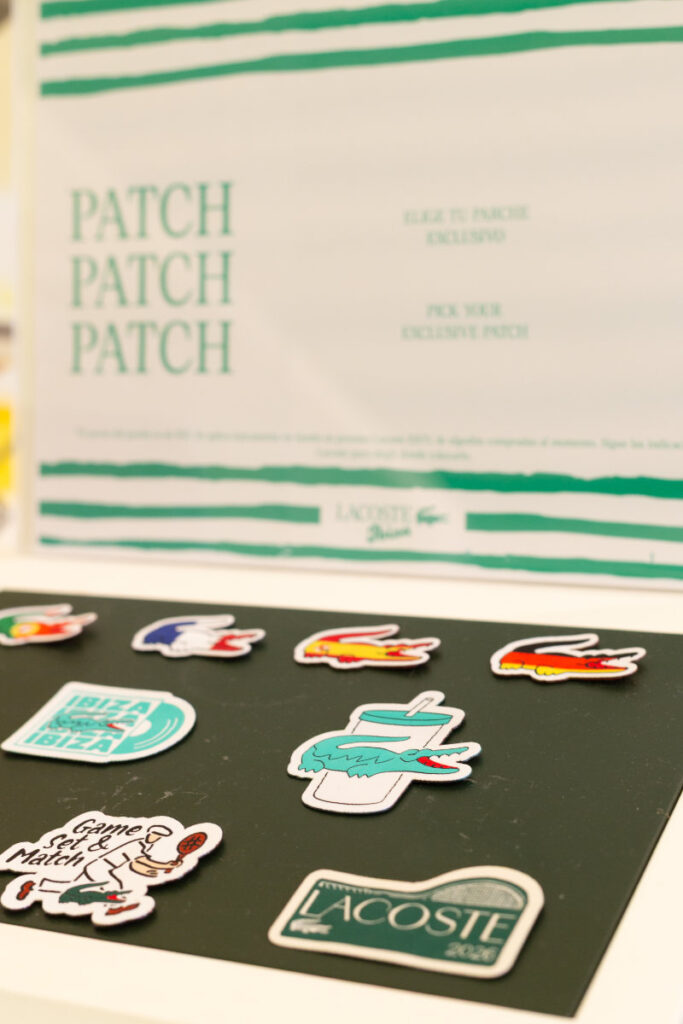 Sticker sheet on a dark surface with various travel-themed icons and logos, including a drink cup and a Lacoste sticker, with a blurred banner reading PATCH PATCH PATCH in the background.