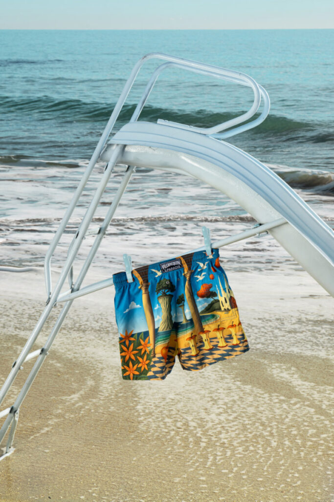 Blue and orange tropical print swim trunks hanging on a lifeguard chair on a sunny beach with the ocean in the background.
