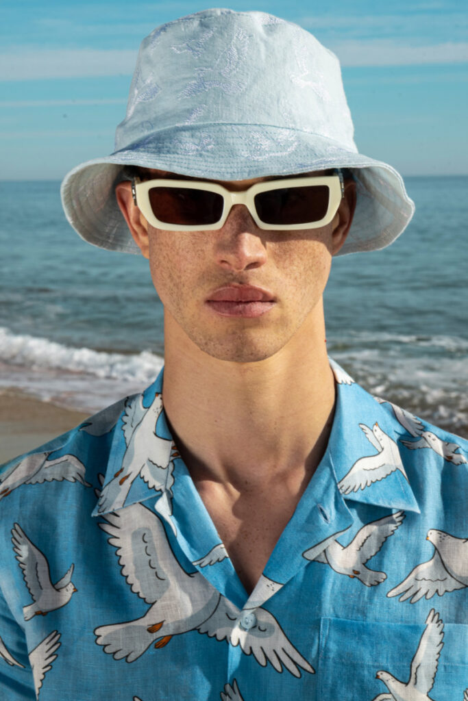 Person on a sunny beach wearing a light blue bucket hat, white rectangular sunglasses, and a blue seagull-print shirt with the ocean in the background.