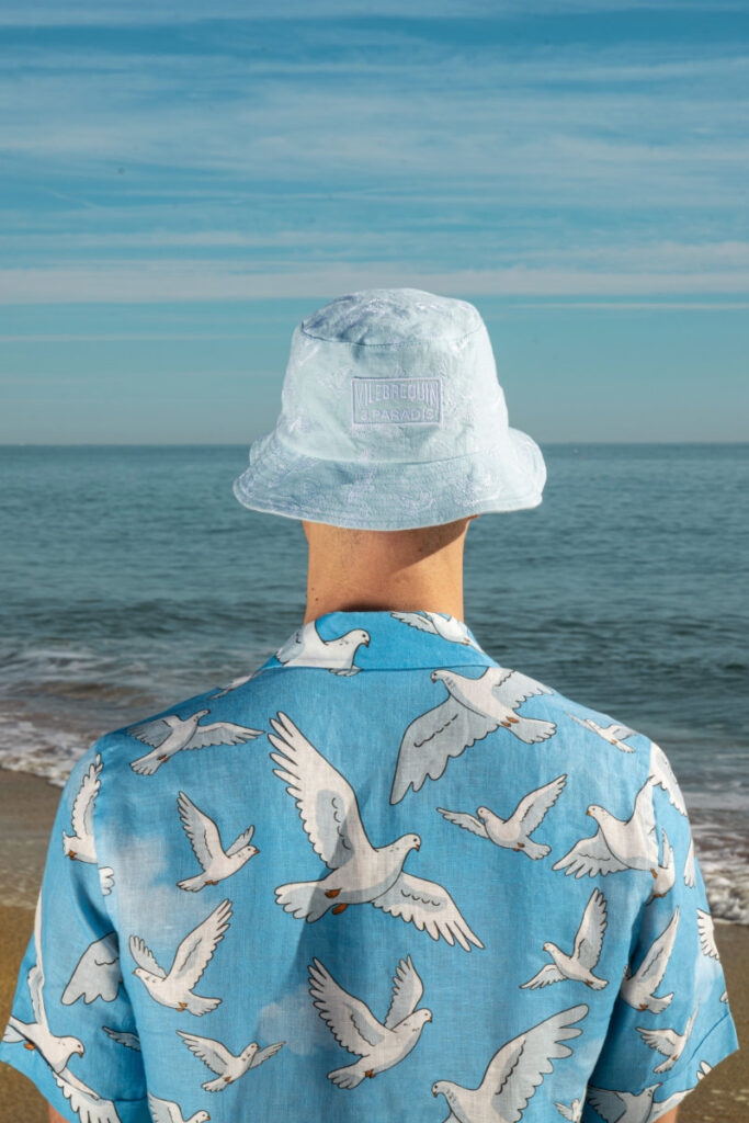 Person standing on a beach, back to viewer, wearing a light blue bucket hat and a blue shirt with white seagull prints, ocean in background.