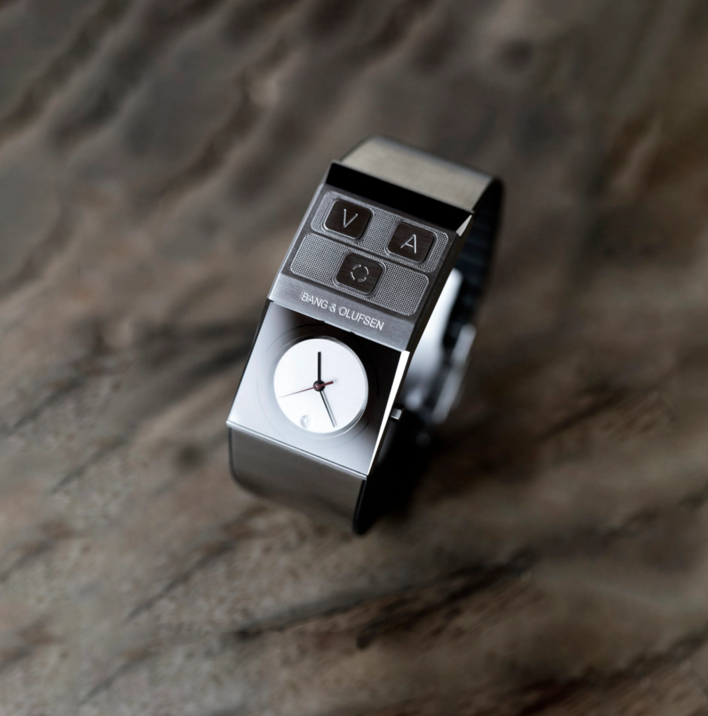Close-up of a Bang & Olufsen device showing metal buttons and a white circular dial (clock-like) at the bottom edge, blurred background