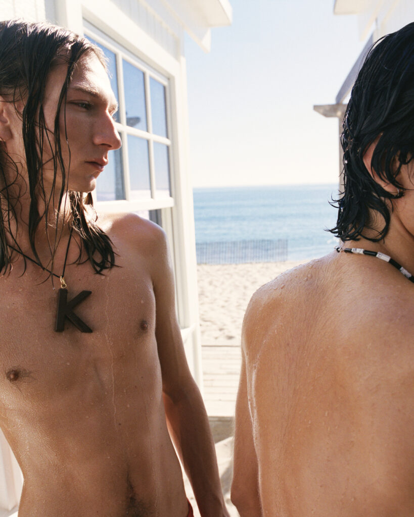 Two shirtless young men on a sunny beach by a white building; one wears a black K pendant.