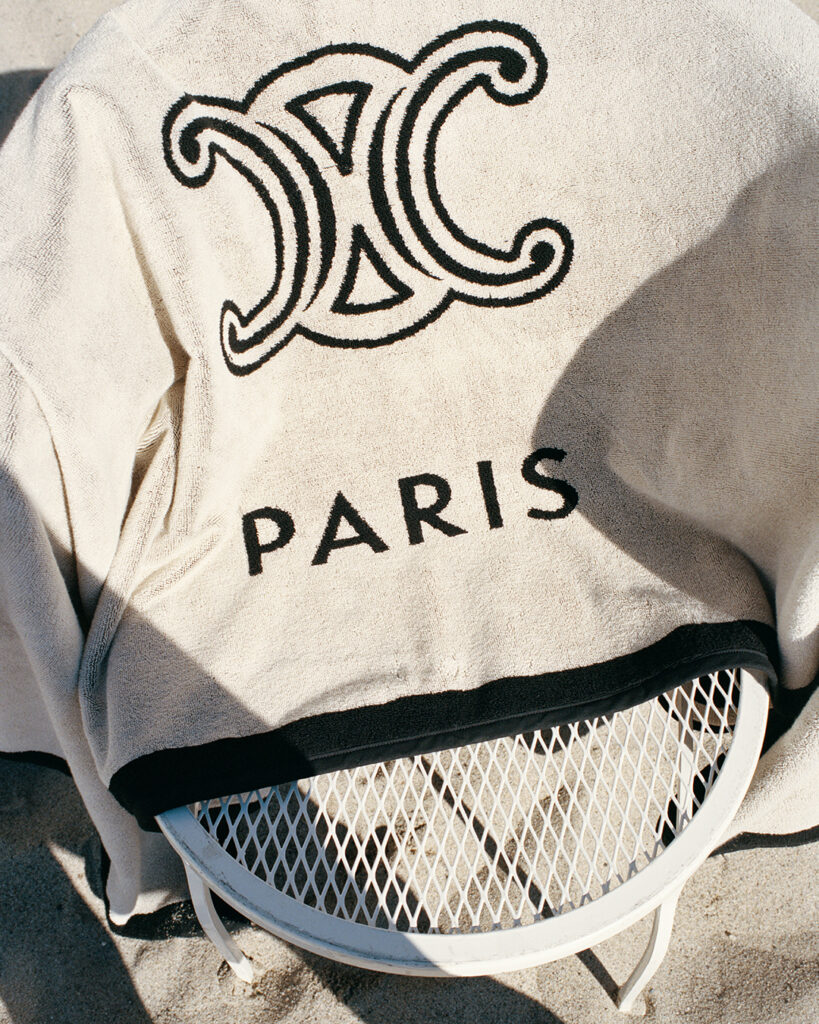 Beige towel with a decorative black logo and the word PARIS draped over a white metal chair on sandy ground in sunlight.",