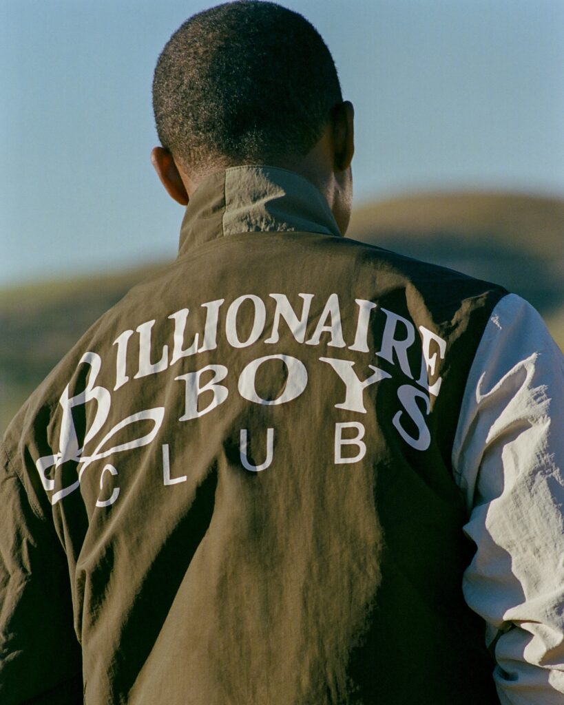 Back view of a person wearing a brown jacket with large white letters reading MILLIONAIRE across the back, outdoors.