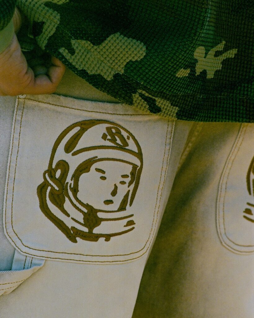 Close-up of white jeans pocket with a brown circular logo featuring a baby’s face, against a camo-pattern fabric background.