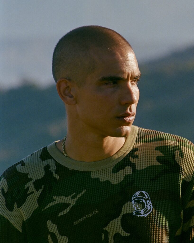 Close-up of a young man with a buzz cut wearing a camo sweater, outdoors with a mountainous background.