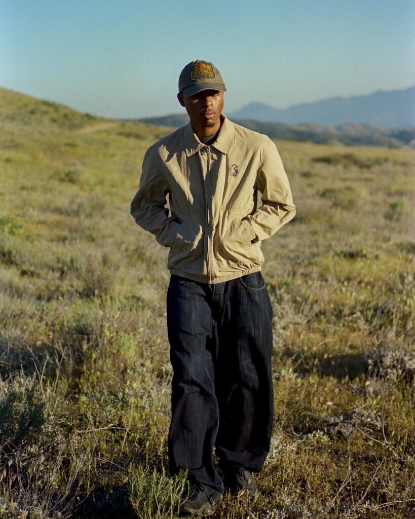 Man in a khaki jacket and cap stands in a dry, grassy field with distant mountains on the horizon.
