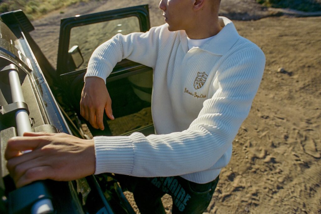 Man in a white ribbed sweater leaning on an open car door in a dusty outdoor setting.