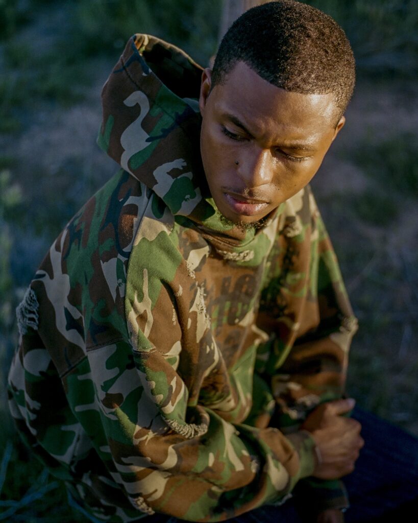 Young man in woodland camouflage jacket looking down, outdoors at dusk with a contemplative expression