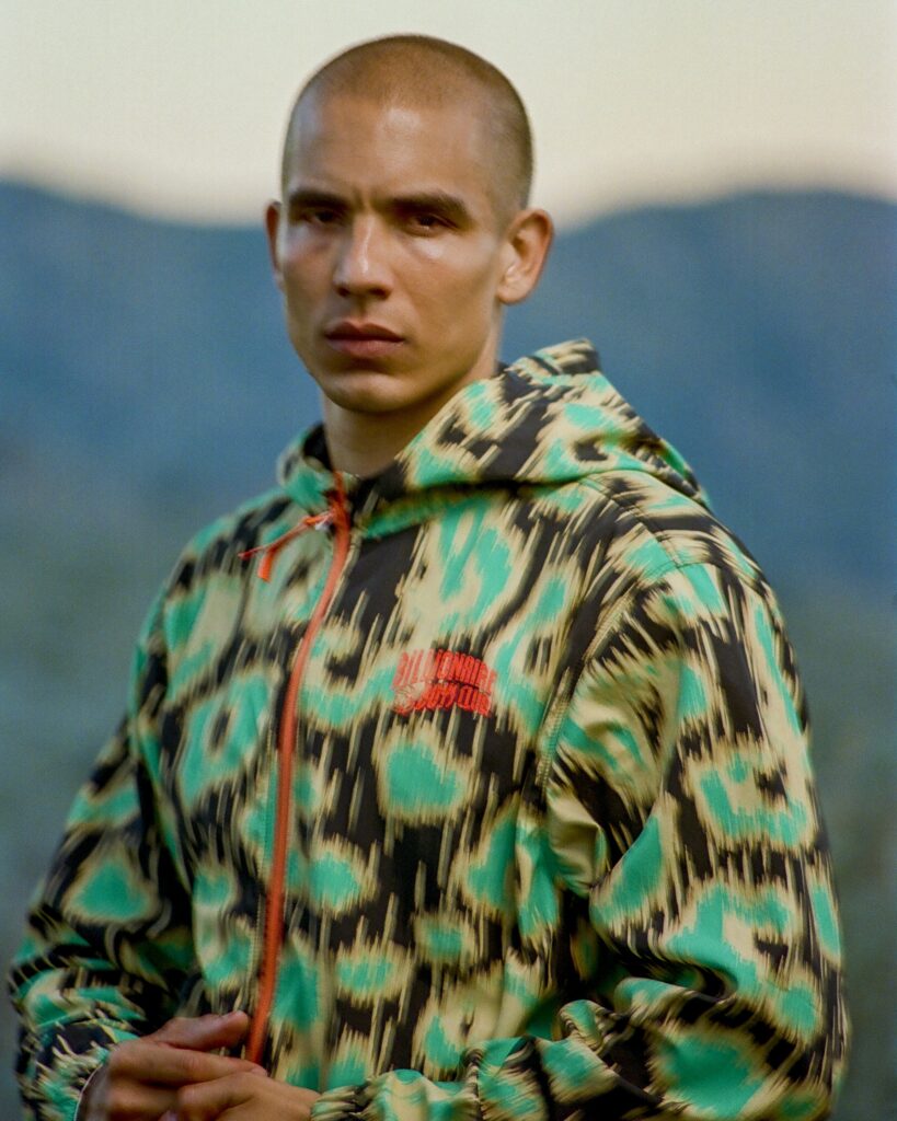 Man with a shaved head wearing a bright camo jacket, standing outdoors with blurred blue mountains in the background.