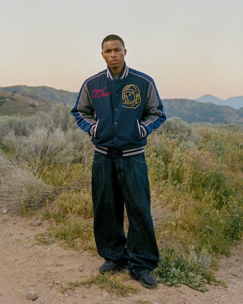 Young man stands in a dry, hilly landscape wearing a blue varsity jacket with patches, facing the camera.