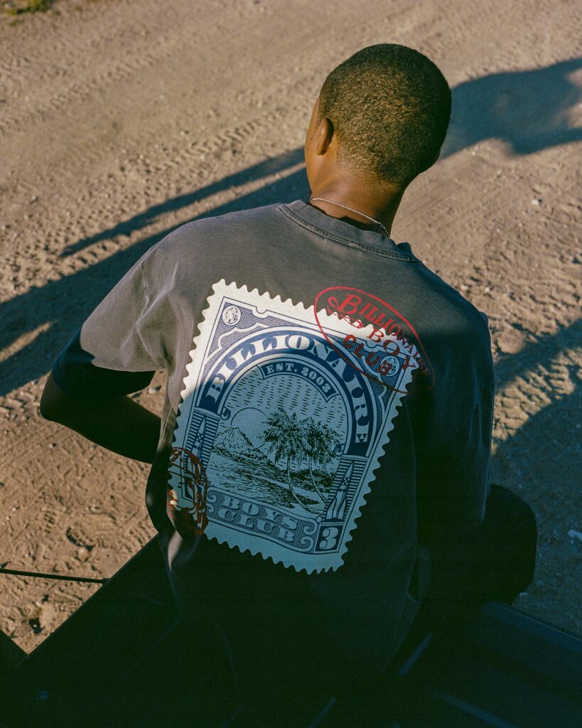 Person viewed from behind wearing a dark gray T-shirt with a large blue stamp-like graphic on the back, outdoors on gravelly ground.