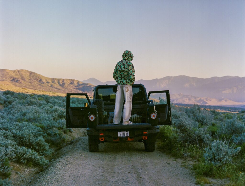 Person wearing a green patterned hoodie standing on the tailgate of a dark SUV in a desert landscape with distant mountains behind them, facing away from the camera.