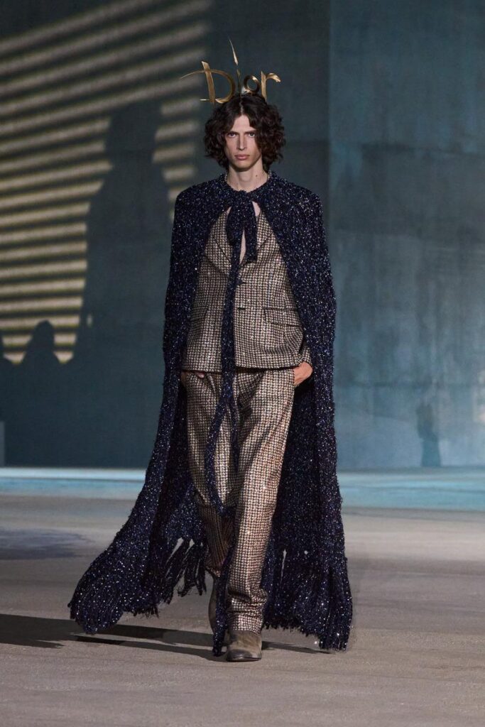 Male model wearing a dark, sequined navy cape over a beige checkered suit with a bow-tied neck at a Dior runway show