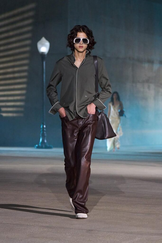 Model walking a fashion runway in an olive shirt with white piping, dark brown leather pants, white sunglasses, and a large dark bag over the shoulder.