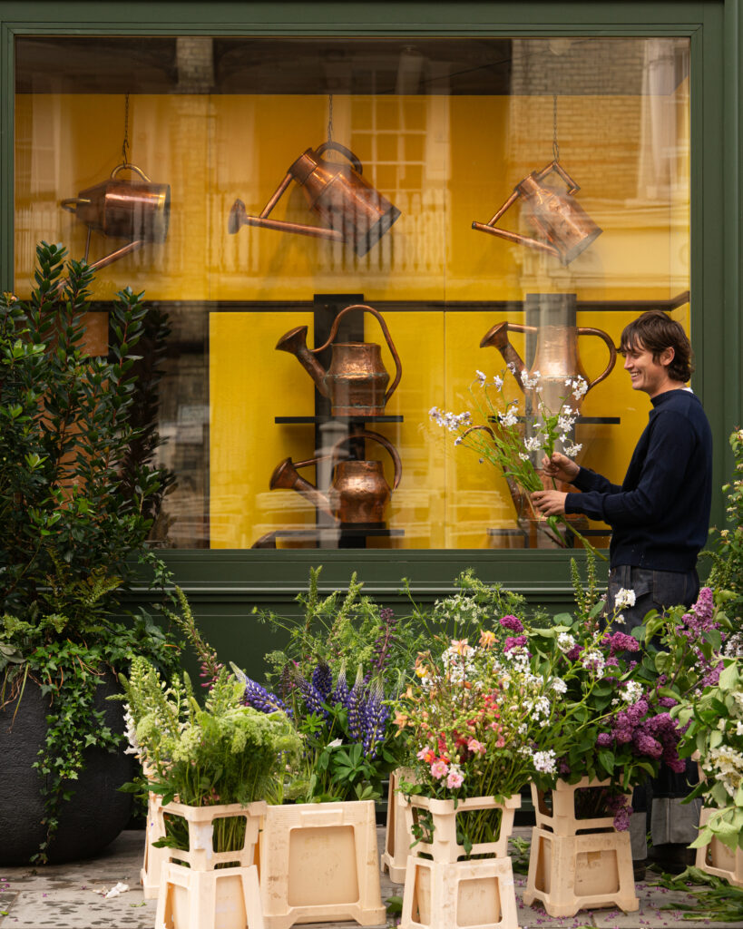 Storefront window display with copper watering cans hanging and potted flowers in crates, a smiling person on the right side of the frame
